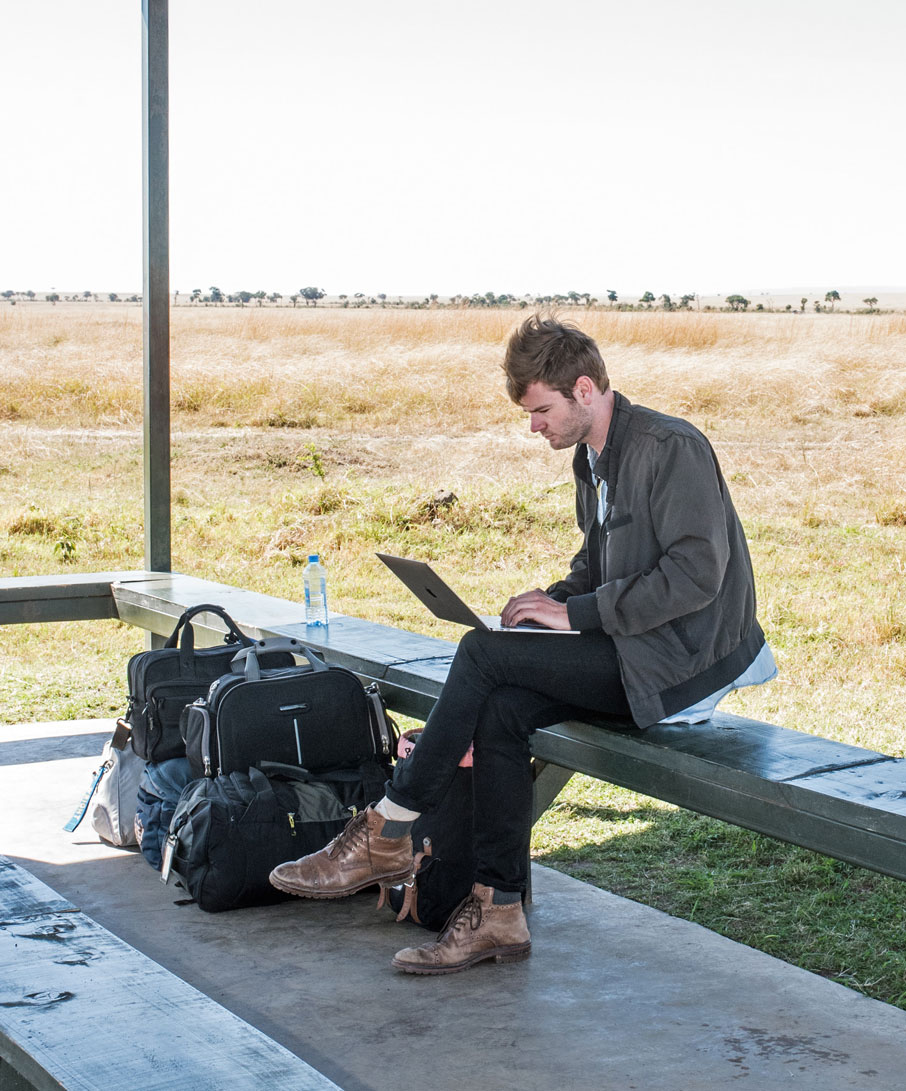 Un jeune homme assis sur un banc de plateforme de train, à l'extérieur, le jour, travaillant sur un ordinateur portable sur les cuisses.
