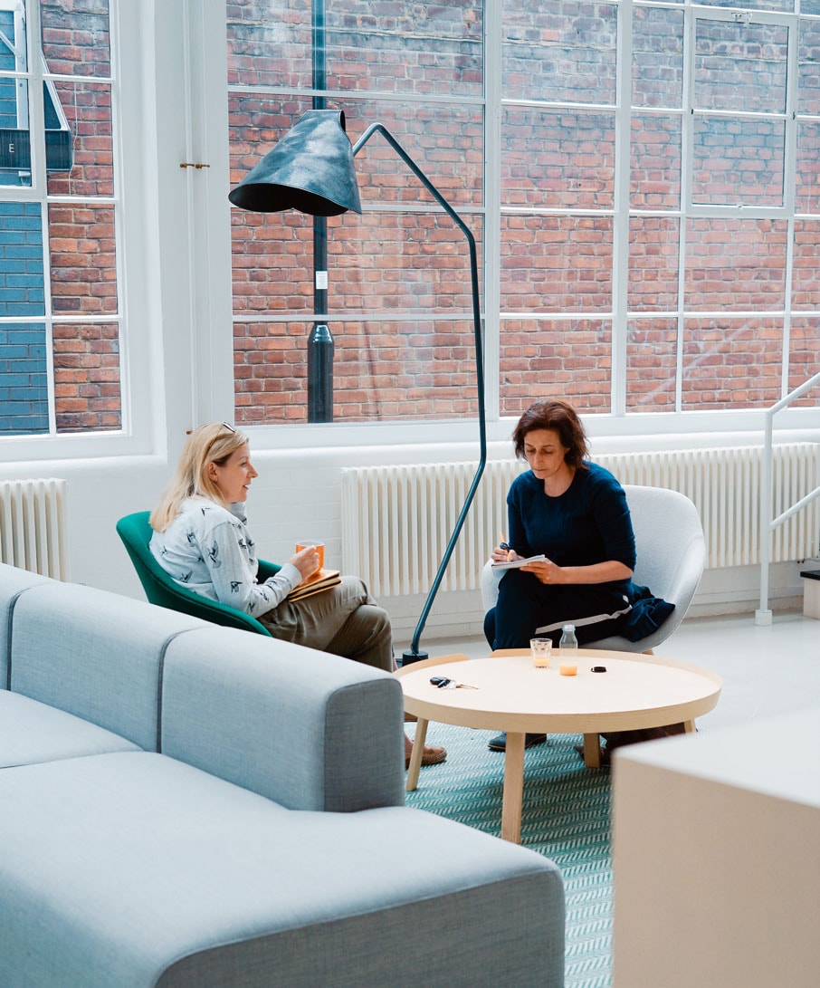 Deux femmes professionnels assis sur des sofas dans un bureau moderne à aire ouvert, en train de discuter.