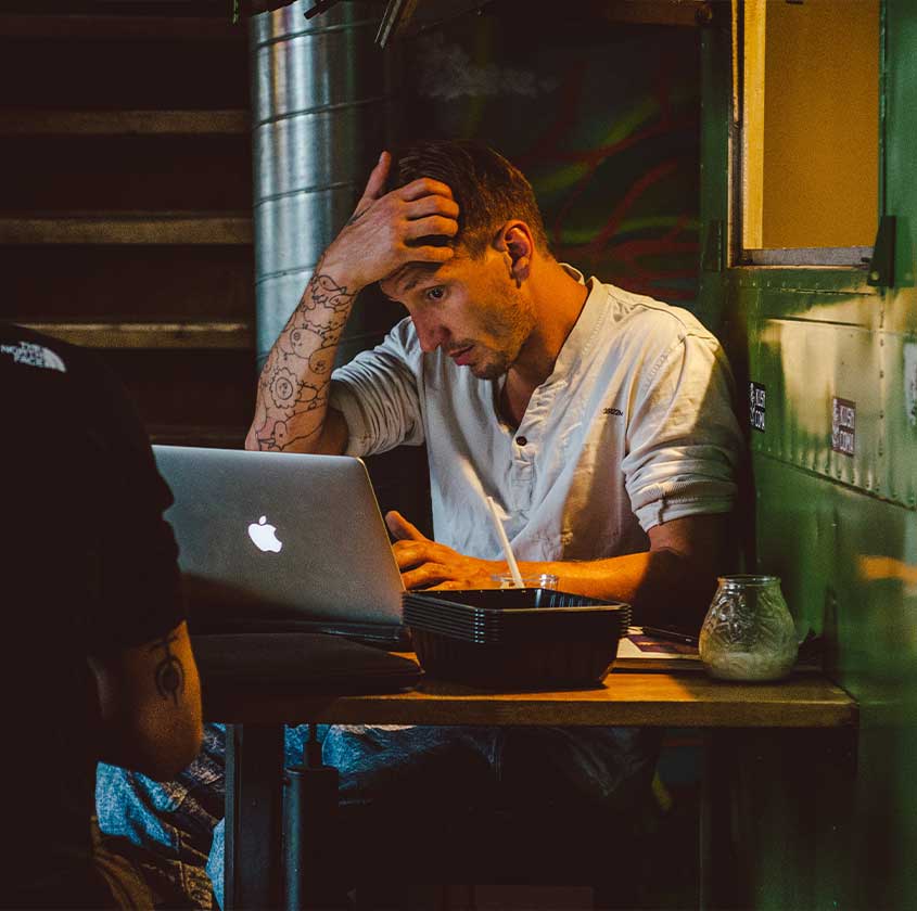 Un homme sur une table de restaurant en bois, la main droite à son front, l'air perplexe, face à son écran d'ordinateur.
